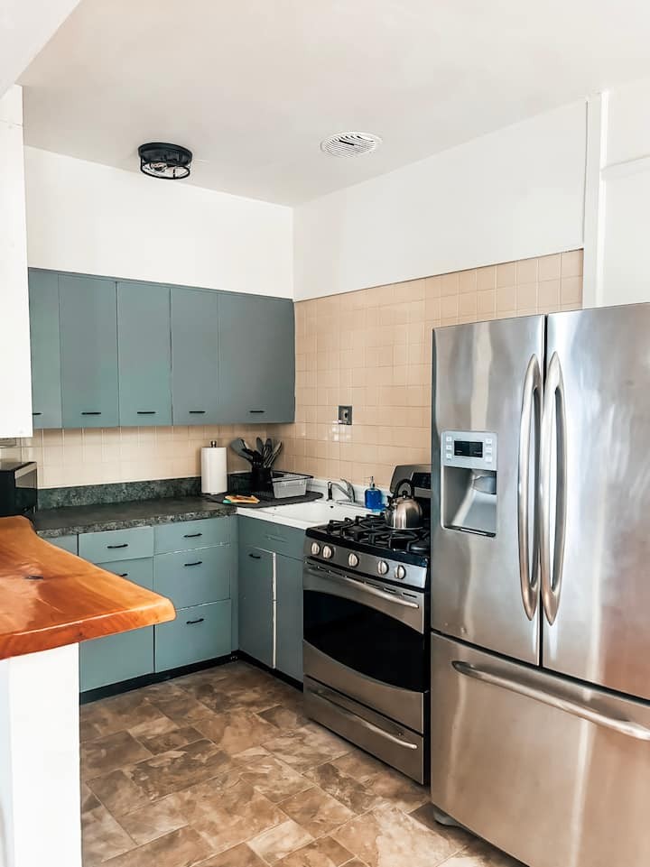 A modern kitchen within 81 Lake Apartments showcasing slate blue cabinetry, stainless steel high-end appliances, and a butcher block island. The backsplash features warm neutral tiles that complement the bright, airy light-mode atmosphere of the boutique residential building.